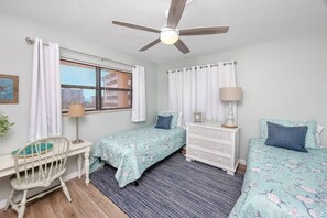 Ein Schlafzimmer mit zwei Betten, einem Schreibtisch, einem Stuhl, einem Deckenventilator und einem Fenster mit Vorhängen.