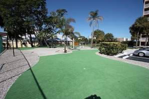 A green artificial turf area with palm trees, a car, and buildings in the background.