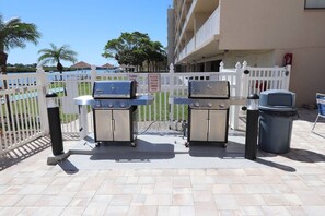 An outdoor barbecue area with two grills, a trash bin, and a white fence.