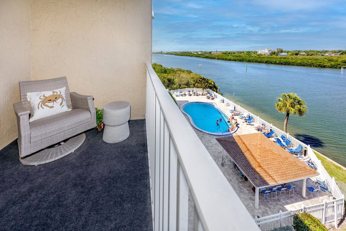A balcony with a view of a river, a pool, and outdoor seating.