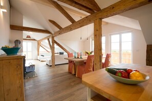A spacious loft with exposed wooden beams, a dining area with a bowl of fruit, and a cozy seating area with a sofa and chairs.