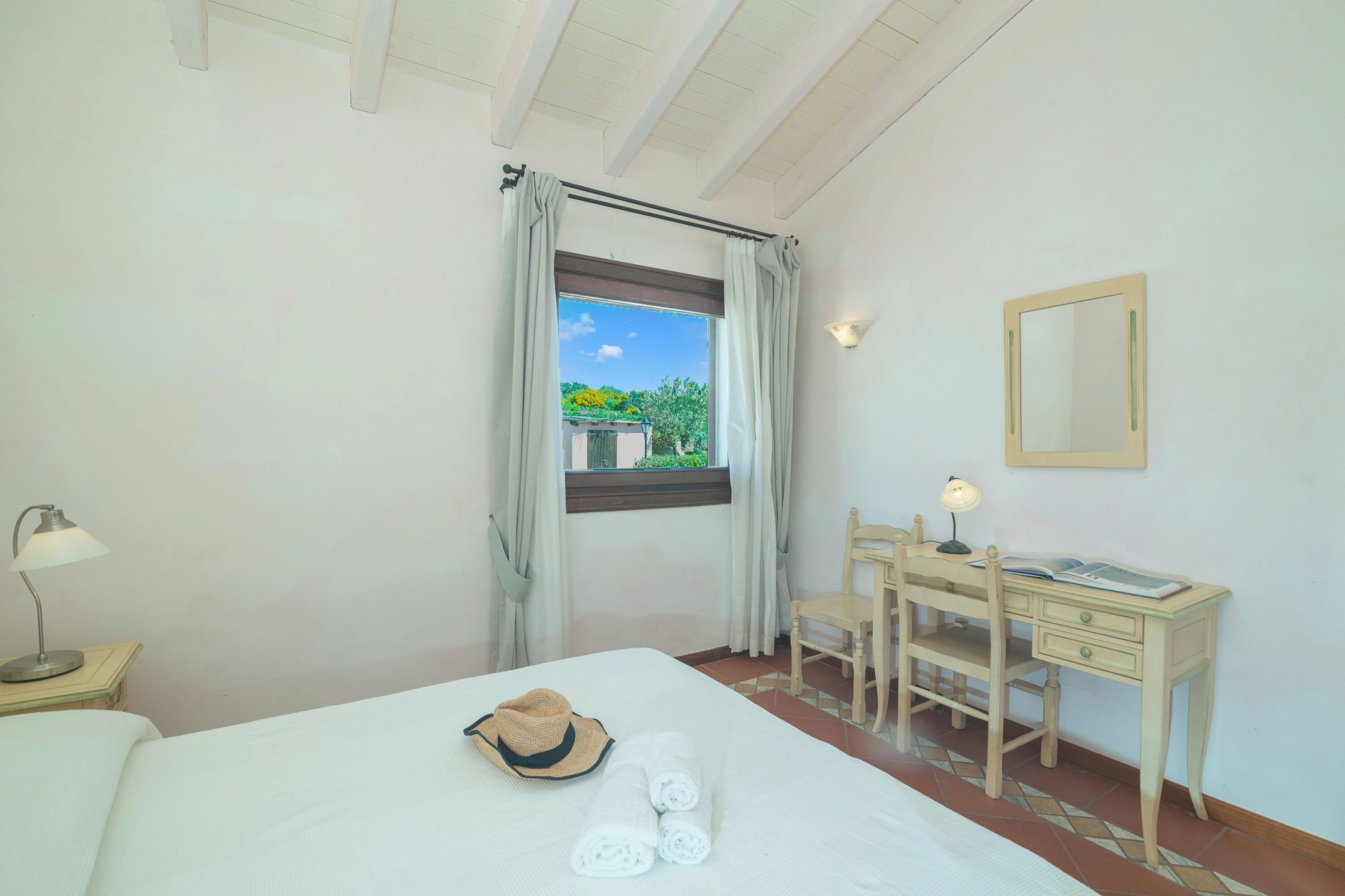 A hotel room with a bed, a desk, a chair, a window with curtains, a lamp, and a hat on the bed.