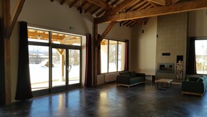 A spacious living area with a high ceiling and exposed wooden beams, large sliding glass doors leading to a snowy outdoor area, a built-in fireplace, and a seating area with a sofa and a small table.