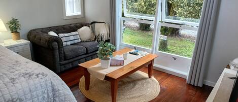 A cozy living room with a dark grey sofa, a wooden coffee table, and a large window offering a view of greenery outside.