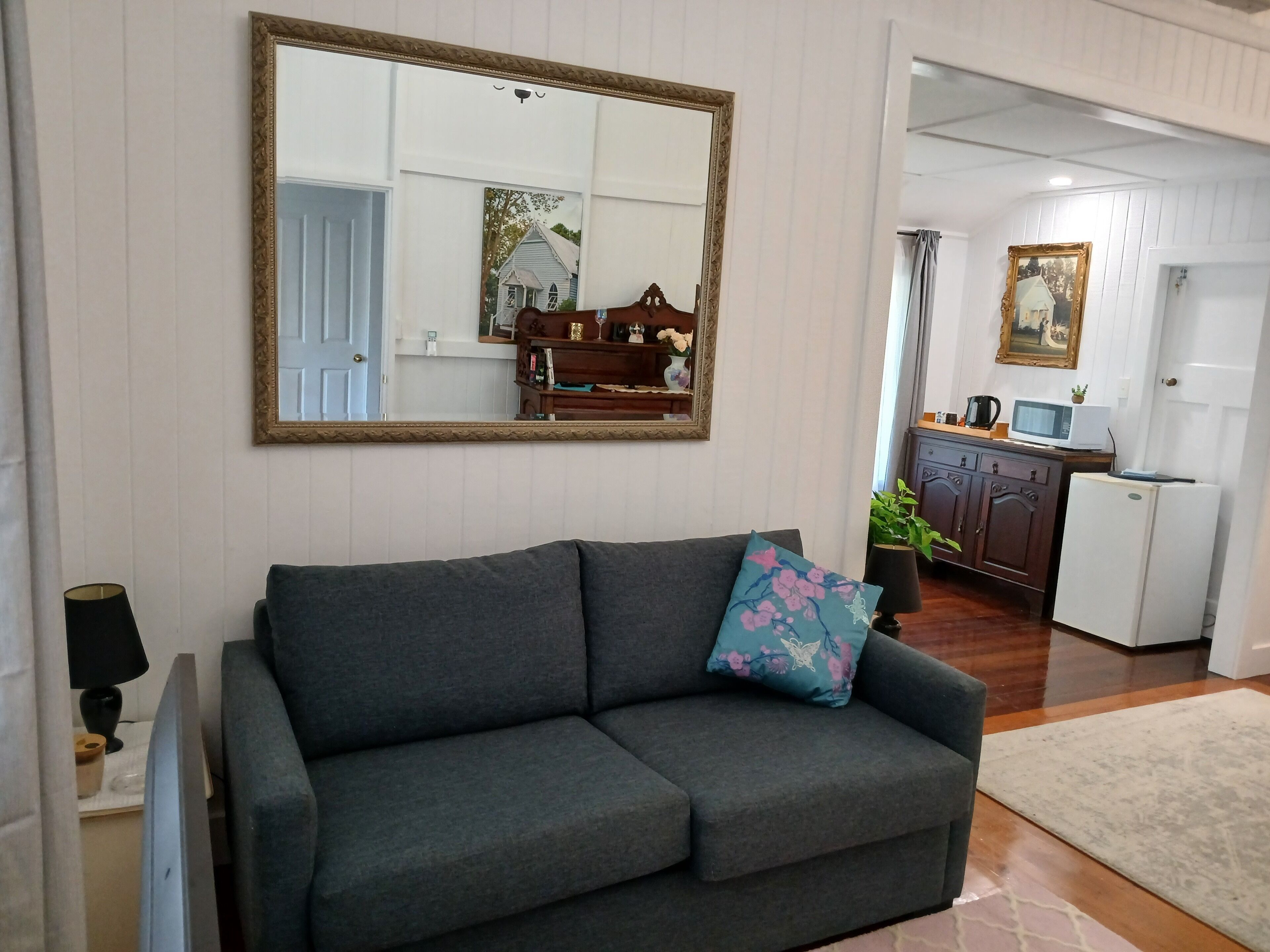 Una sala de estar con un sofá gris, un espejo grande y una cocina al fondo.