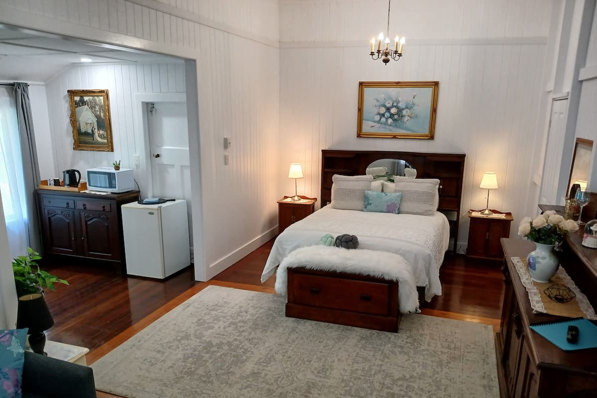A bedroom with a bed, bedside tables, a dresser, a desk, a chair, a small kitchenette, and a door leading to another room.