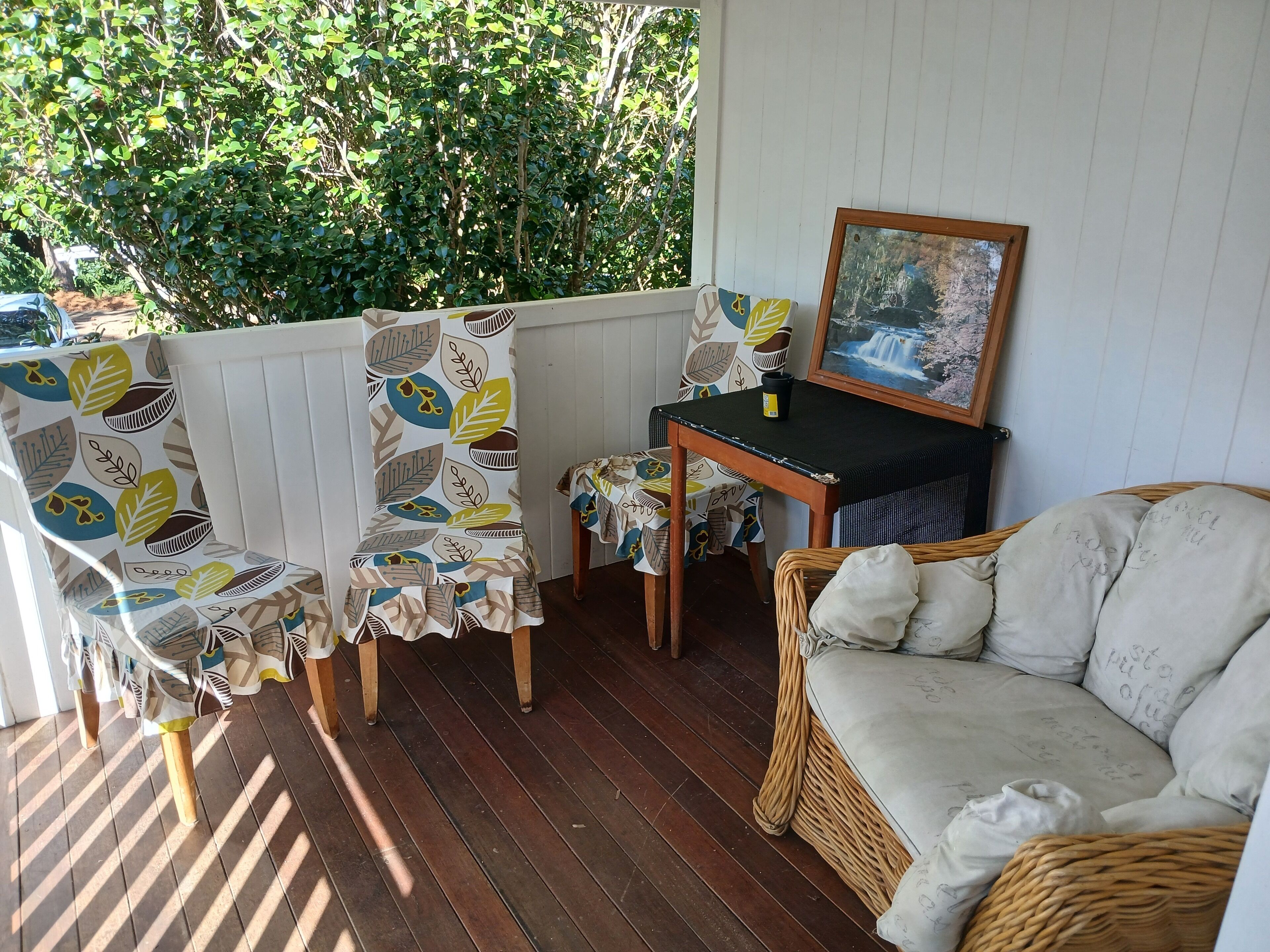 A covered outdoor seating area with a wicker sofa, chairs with leaf-patterned upholstery, a small table with a picture frame, and a view of greenery through the window.