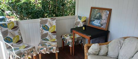 A covered outdoor seating area with a wicker sofa, chairs with leaf-patterned upholstery, a small table with a picture frame, and a view of greenery through the window.