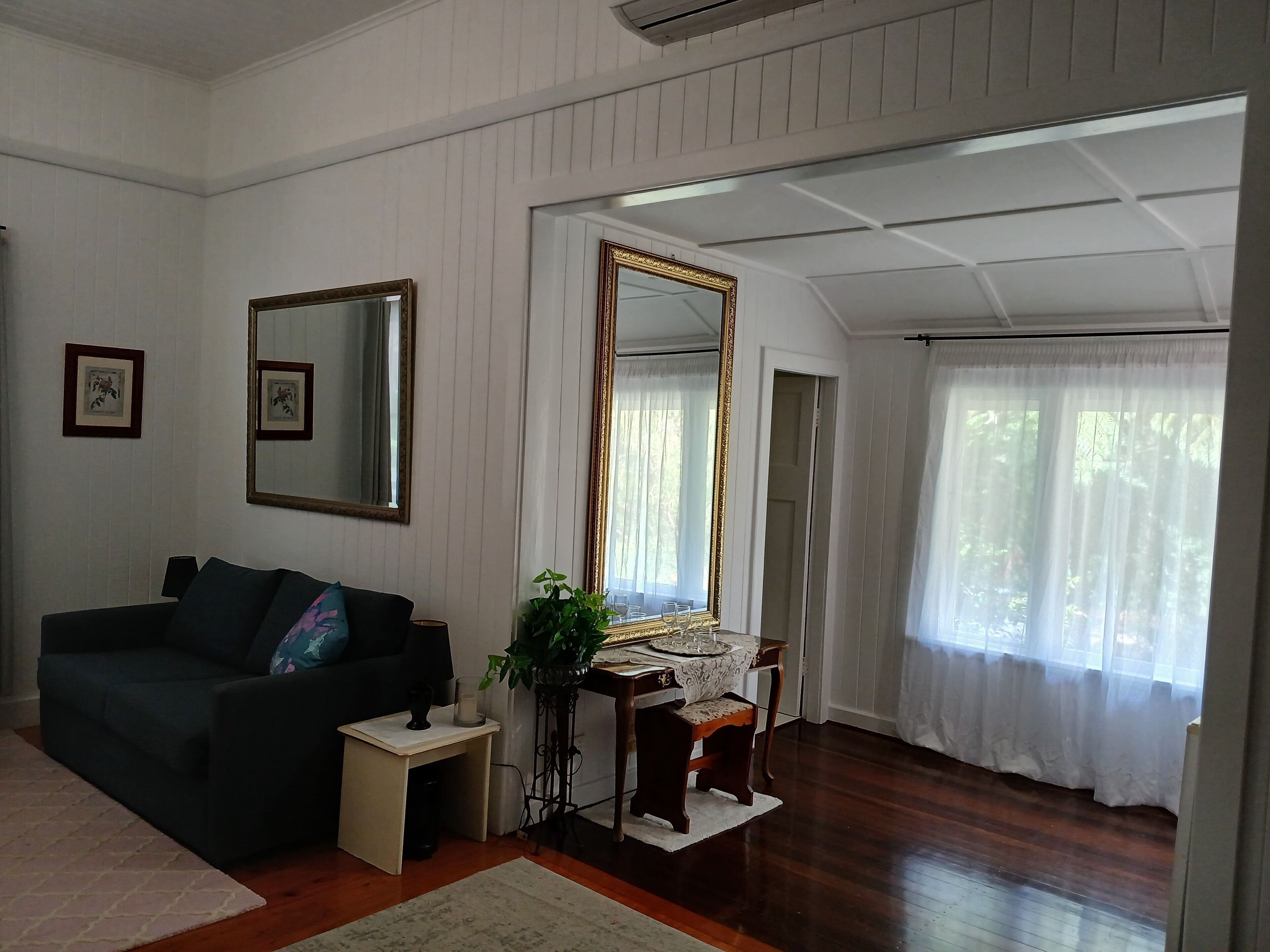 A living room with a dark-colored sofa, a small side table, a mirror, and a dining table with a lace tablecloth.