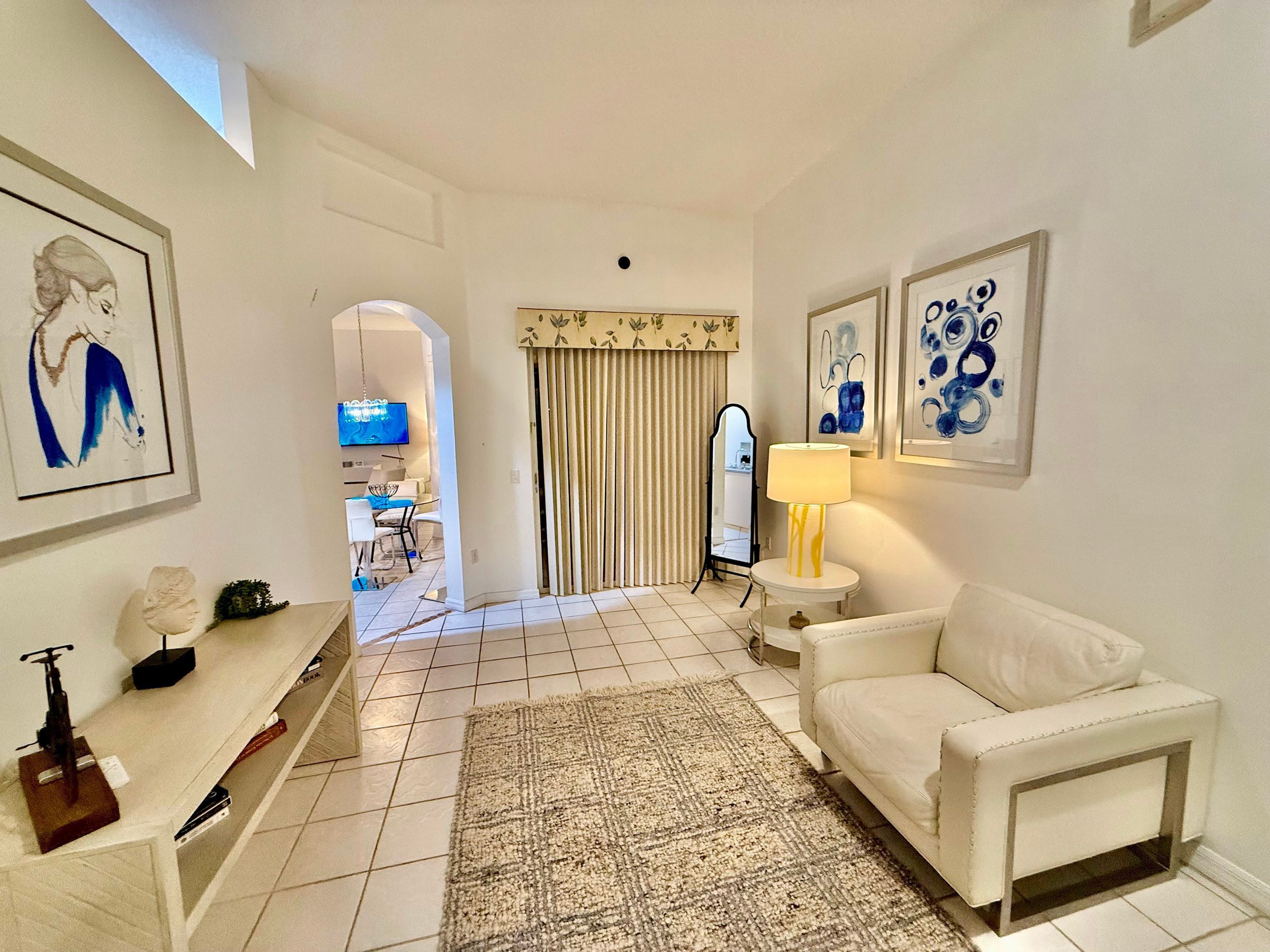 A well-lit living room with a white sofa, a small round table with a lamp, a TV, and a dining area in the background.