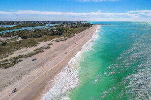 An aerial view of a beach with clear turquoise waters, a sandy shoreline, and a backdrop of greenery and buildings.