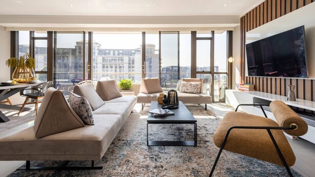A modern living room with a sofa, armchairs, a coffee table, and a TV. Large windows provide a view of the cityscape. - Azalia Apartments (Sandton)