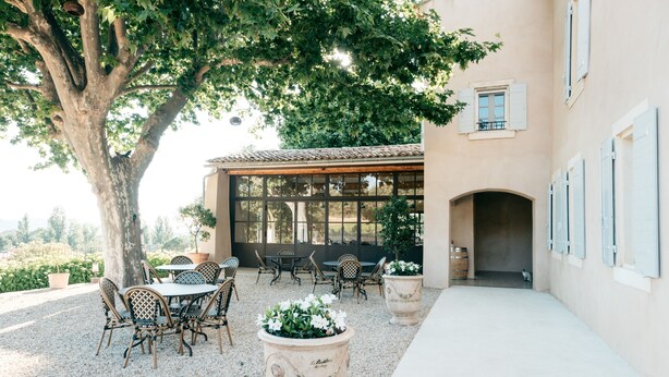 Un patio al aire libre con mesas y sillas, un árbol grande y un edificio con ventanas y persianas.