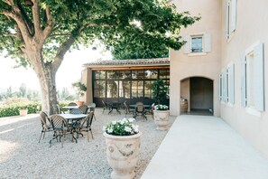 An outdoor patio with tables and chairs, a large tree, and a building with windows and shutters. - Domaine Perréal (Gargas)
