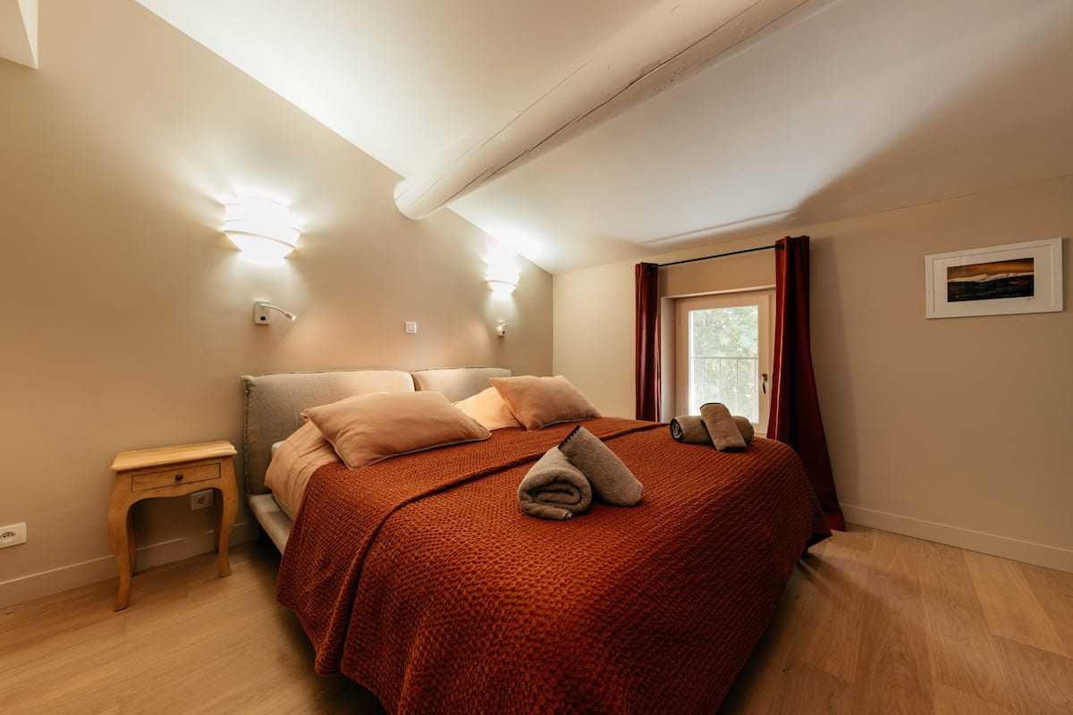 Quarto com cama, mesa de cabeceira, luminária na parede e janela com cortinas.