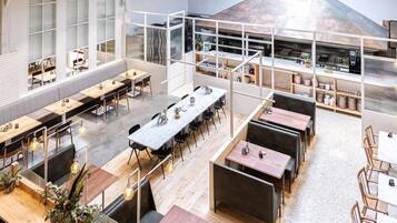 Un vaste espace dédié à la salle à manger, avec de nombreuses tables et des bancs, de grandes fenêtres et un coin cuisine visible à travers une paroi en verre.