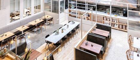 A spacious dining area with multiple tables and benches, large windows, and a kitchen area visible through a glass partition.