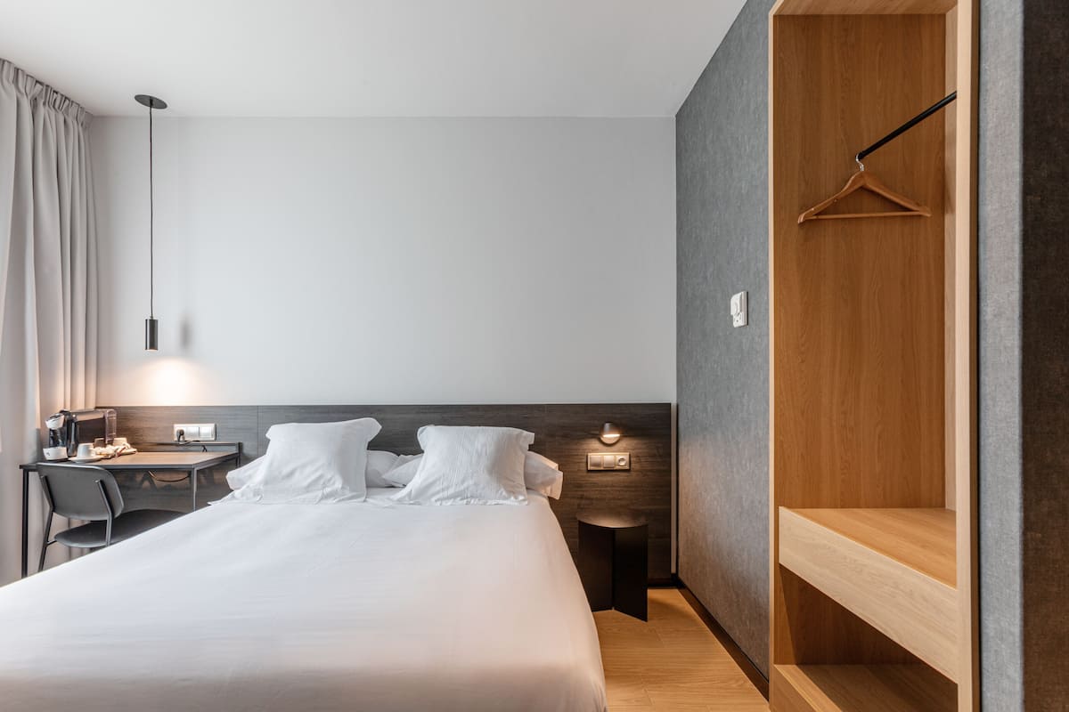 A modern hotel room with a large bed, a desk with a chair, a hanging light fixture, and a wooden wardrobe.