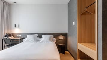 A modern hotel room with a large bed, a desk with a chair, a hanging light fixture, and a wooden wardrobe.