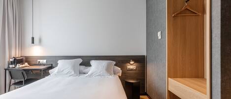 A modern hotel room with a large bed, a desk with a chair, a hanging light fixture, and a wooden wardrobe.