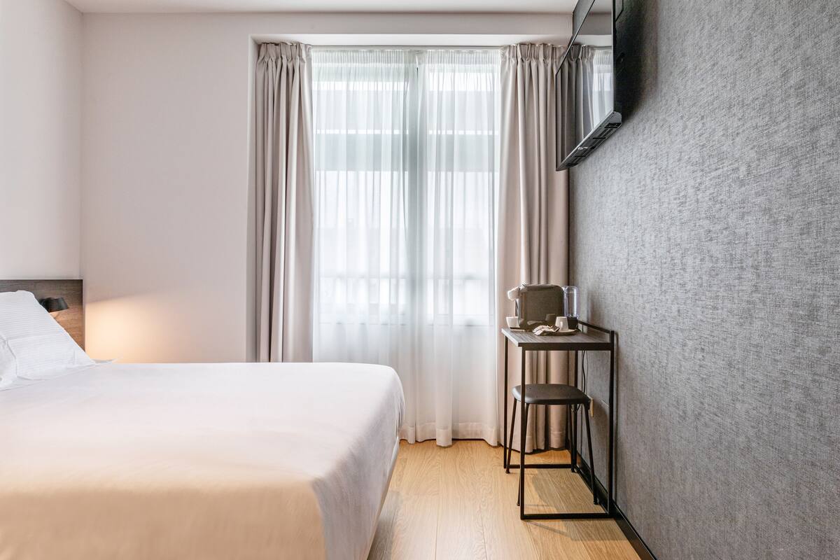 A hotel room with a bed, a desk with a chair, a flat-screen TV mounted on the wall, and a large window with sheer curtains.