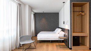 Una camera d'albergo moderna con un letto, una sedia, un tavolino, un armadio e uno specchio.
