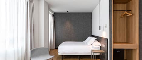 A modern hotel room with a bed, a chair, a small table, a wardrobe, and a mirror.