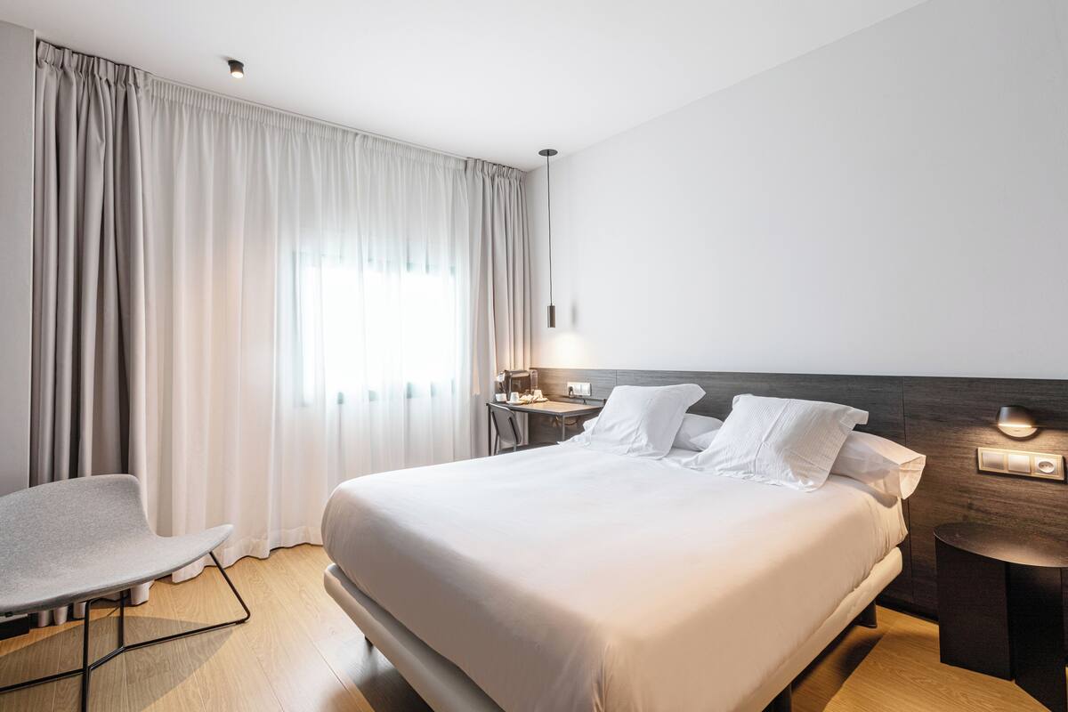A modern hotel room with a large bed, a desk, a chair, and a nightstand with a lamp.