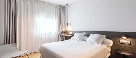 A modern hotel room with a large bed, a desk, a chair, and a nightstand with a lamp.