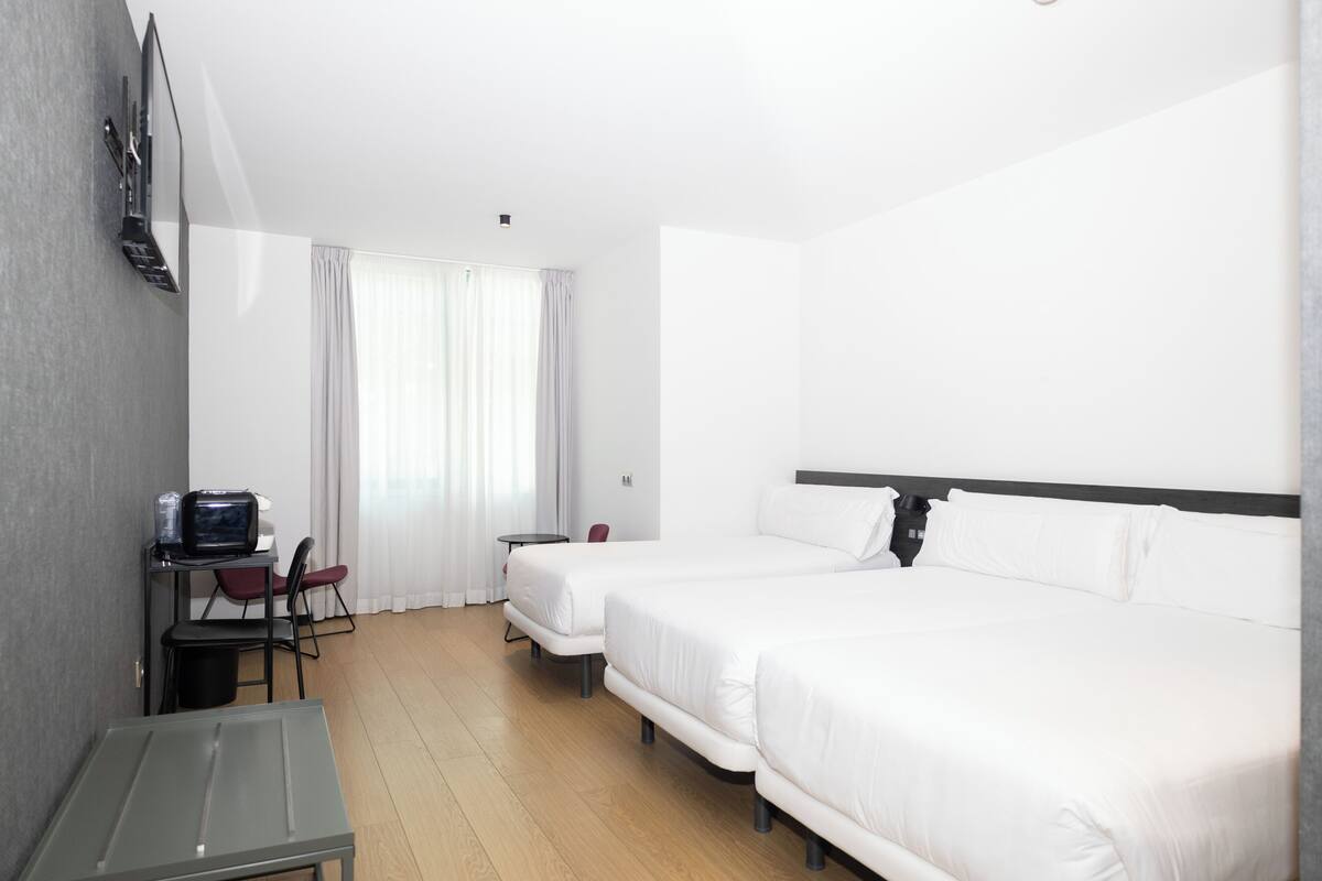 A hotel room with two single beds, a desk with a chair, a small table, and a TV mounted on the wall.