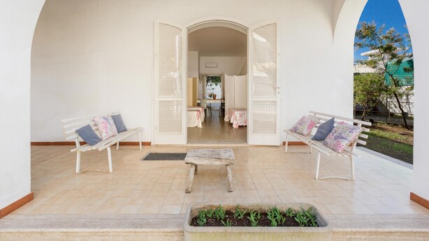 A covered outdoor area with two white benches, a small table, and a view into a room with a table and chairs. - 3821 VBR - Mono Rosa by Barbarhouse (Porto Cesareo)