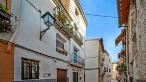 A narrow street with buildings on both sides, featuring balconies with plants, a wooden door, and a tiled ground. - 7 Pisos Casa Rural de Pueblo (Cocentaina)
