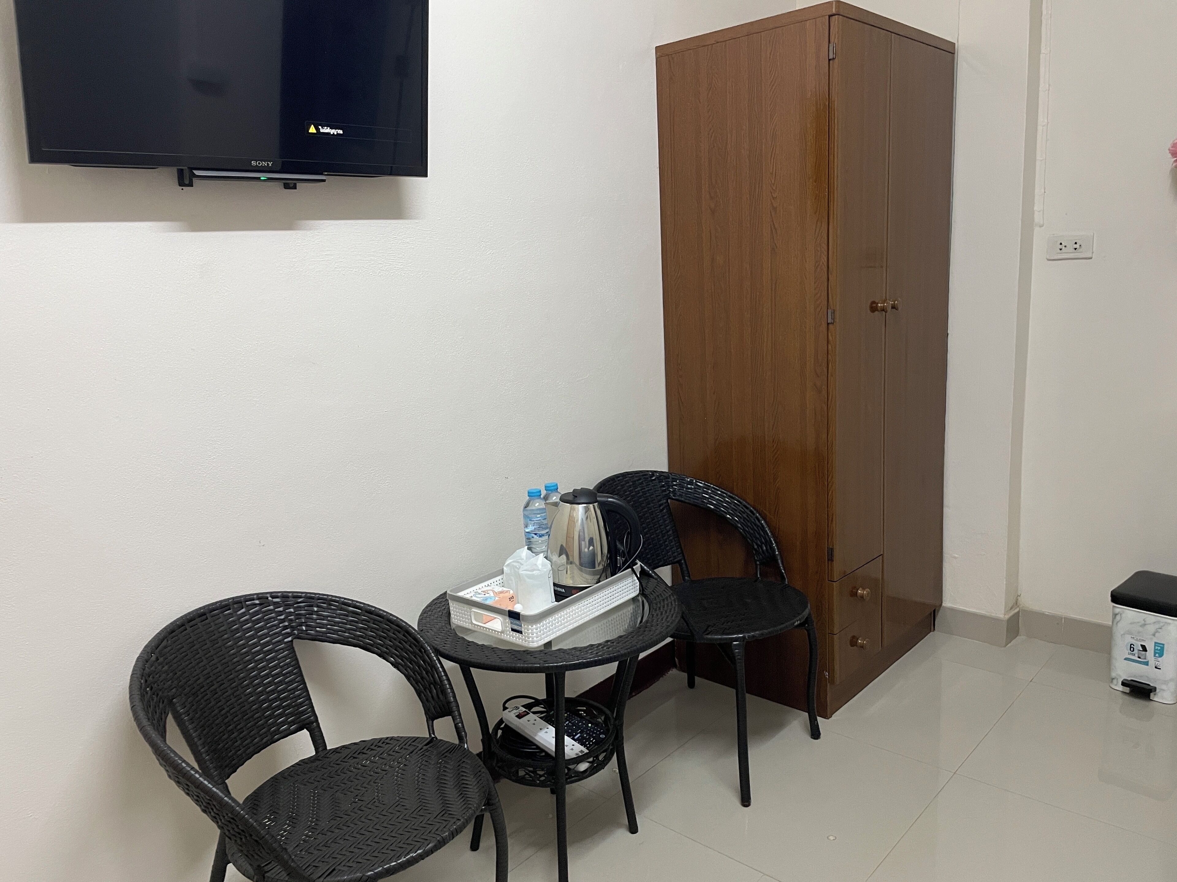 Una habitación pequeña con televisión, un armario de madera, dos sillas de mimbre, una mesa con una botella de agua y una tetera, y un cubo de basura.