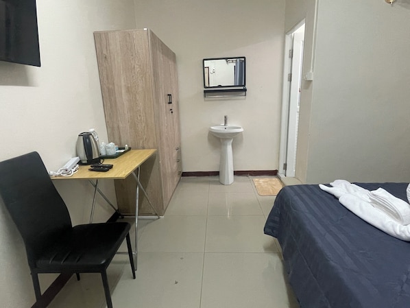A compact hotel room with a bed, a desk with a chair, a wardrobe, a sink, and a mirror.