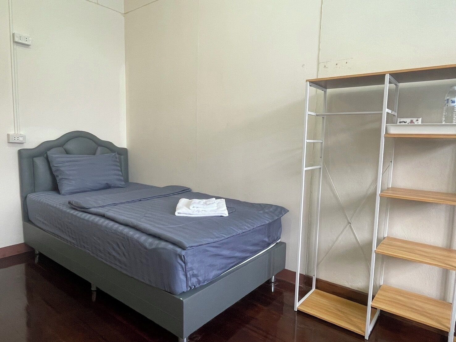 A single bed with a grey headboard and pillows, a folded white towel on the bed, and a metal shelf unit with wooden shelves to the right.