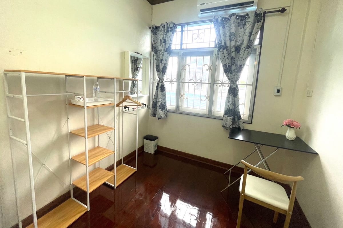 A room with a wooden floor, a metal wardrobe, a small table with a vase, and a window with curtains.