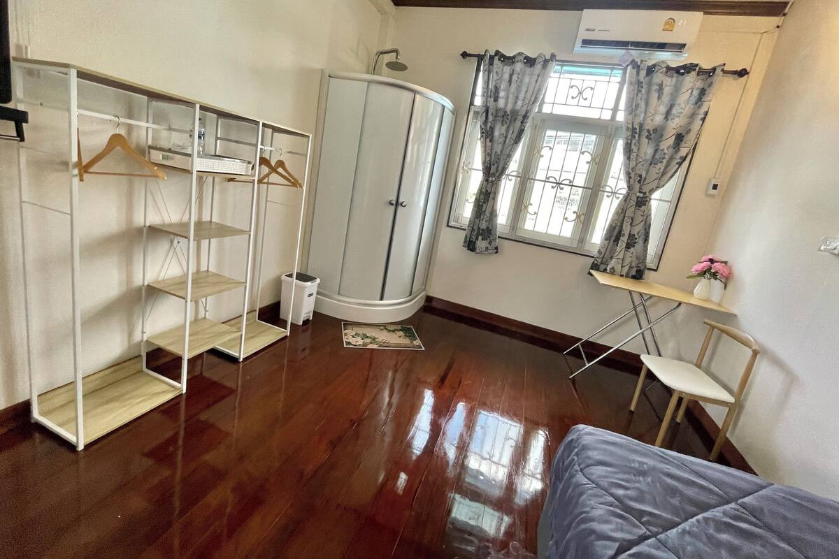 A room with a wardrobe, a bed, a small table with a chair, and a window with curtains.