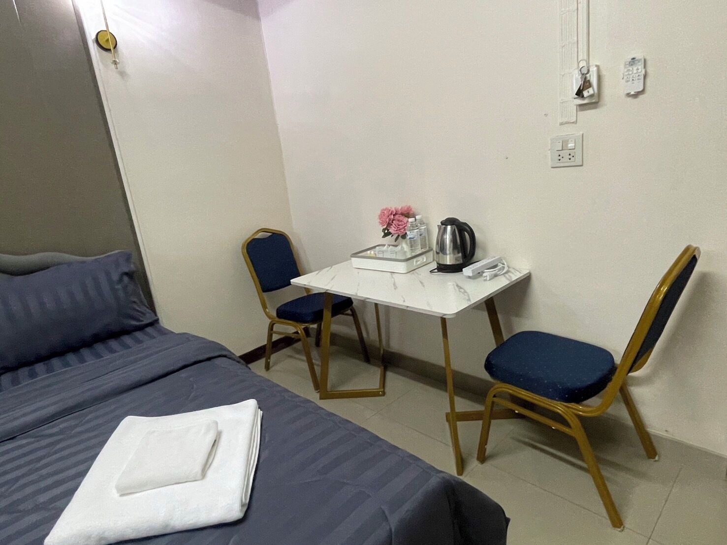 Une chambre d’hôtel avec un lit, deux chaises, une petite table et un bureau équipé d’une bouilloire et agrémenté de fleurs.