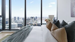 A high-rise hotel room with a large window offering a city view, a bed with multiple pillows, and a lamp on the nightstand.