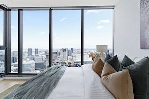 A high-rise hotel room with a large window offering a city view, a bed with multiple pillows, and a lamp on the nightstand. - A Cozy 2BR Apt with Harbour Views, FREE Parking (Melbourne)