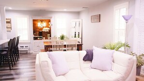 Una sala de estar moderna con un sofá blanco, una zona de comedor con una mesa y sillas de madera, y una cocina con una barra para café.