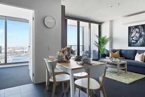 A modern living room with a dining area, large windows, a city view, a clock, and a painting on the wall.