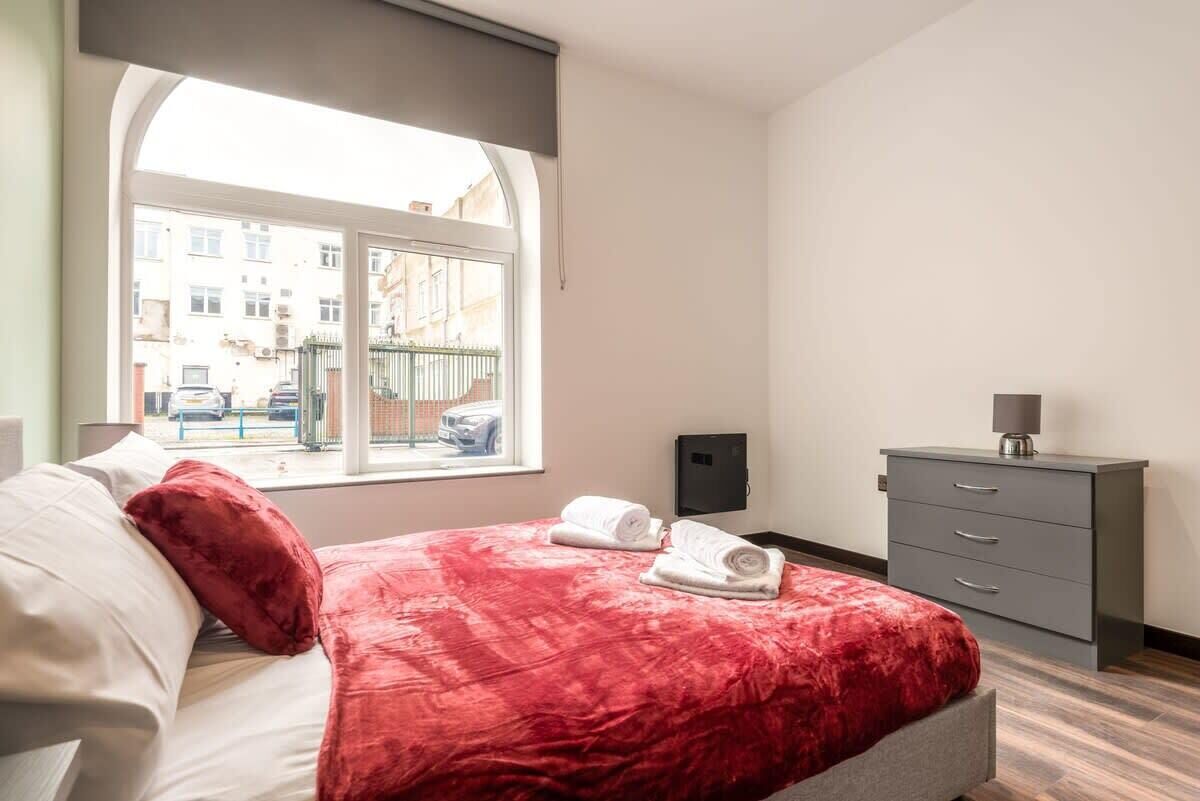A hotel room with a large window, a bed with red bedding, a grey dresser, and a wall-mounted TV.