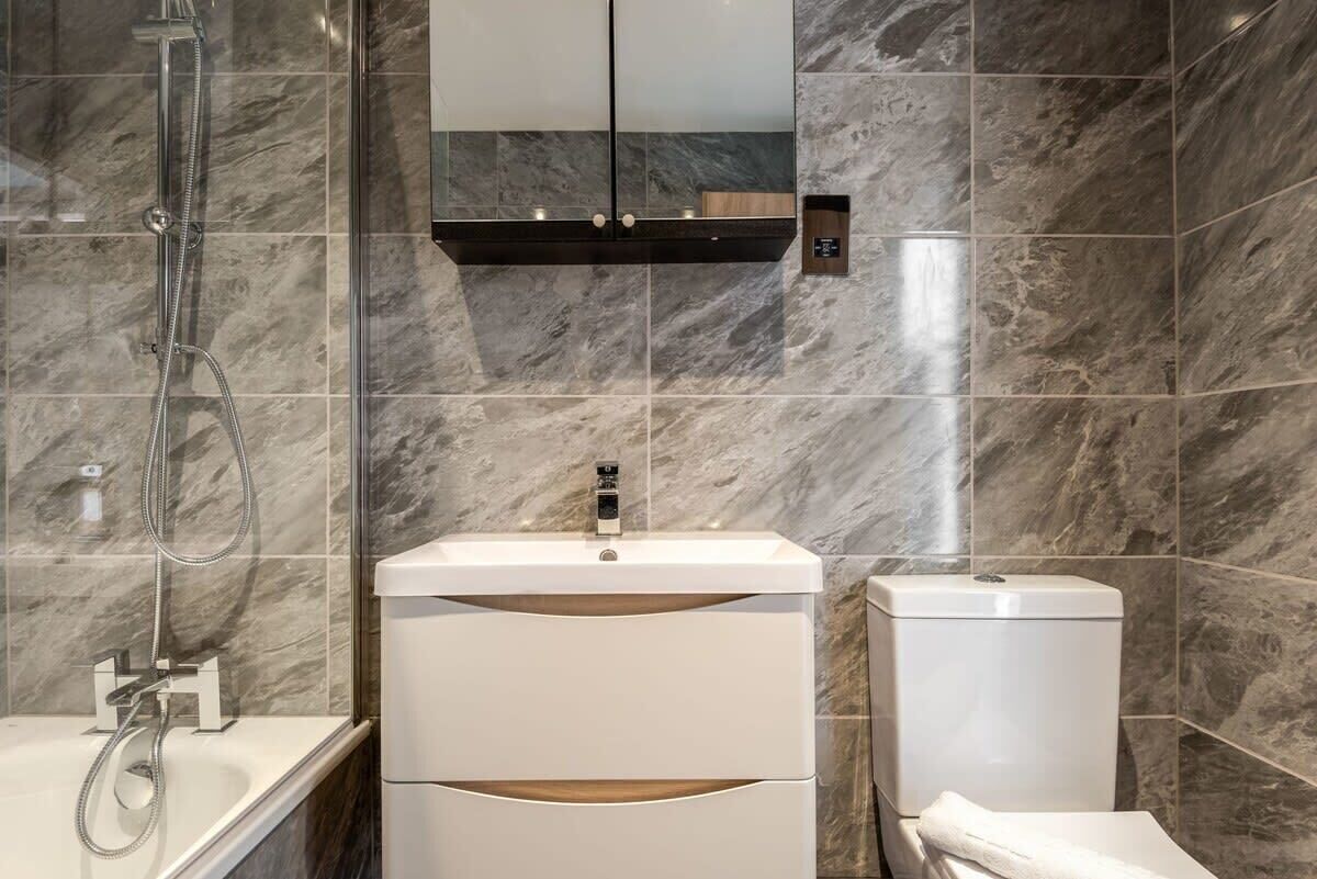 Une salle de bain moderne avec un mur recouvert de carreaux de marbre, un lavabo blanc avec un miroir au-dessus, une douche et des toilettes.