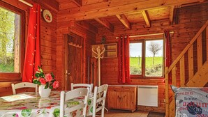 Interno di una baita in legno con un tavolo da pranzo, sedie, un vaso di fiori, una sveglia e una finestra che si affaccia su un prato verde.