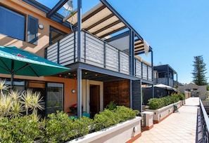 A modern building with a covered outdoor seating area, a balcony, and a pathway leading to a gated entrance.