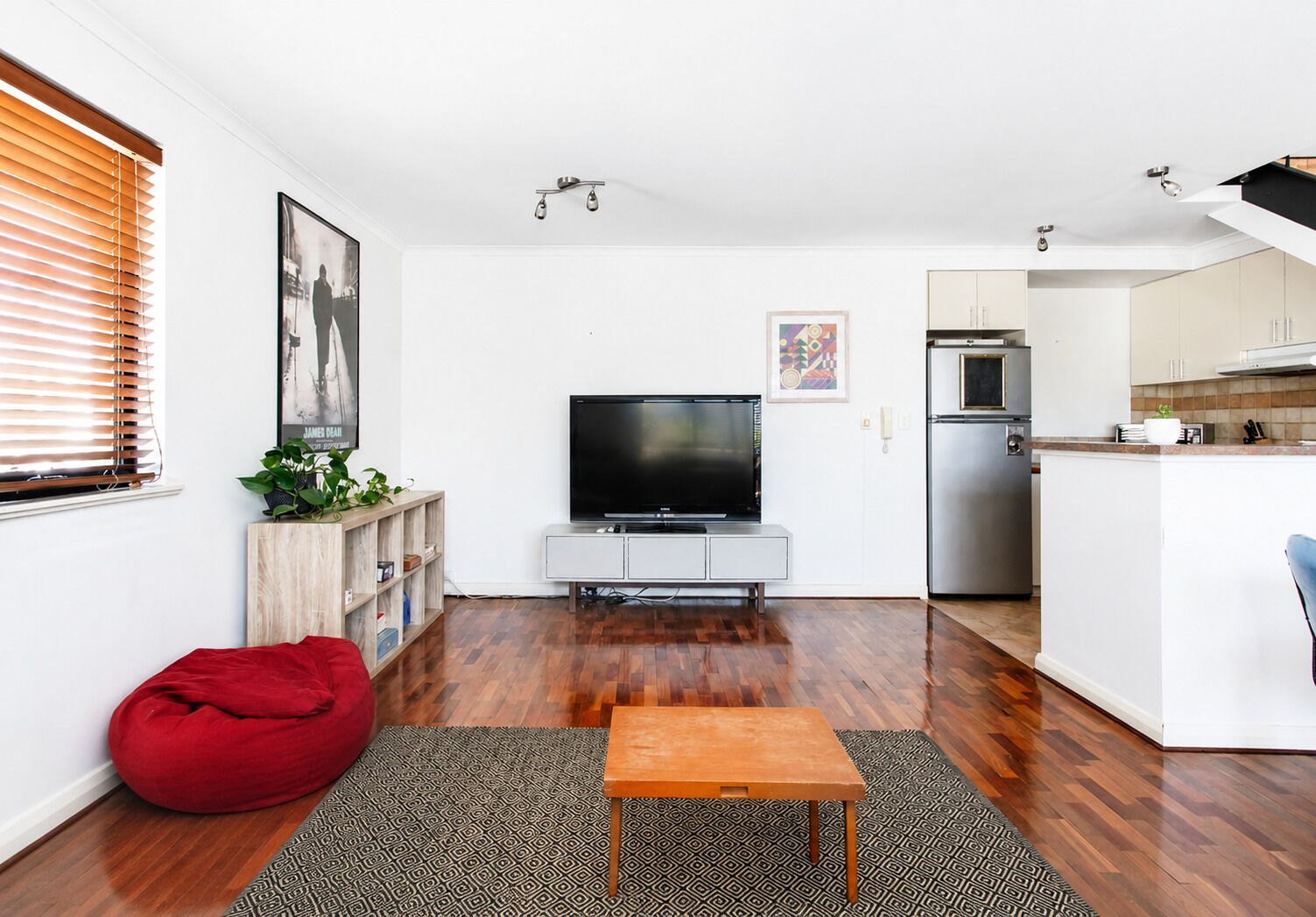A modern living room with a flat-screen TV, wooden flooring, a red bean bag, and a kitchen with a built-in fridge and microwave.