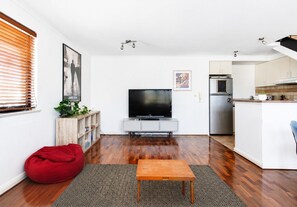 Un salon moderne doté d’un téléviseur à écran plat, de parquet, d’un fauteuil poire rouge et d’une cuisine équipée d’un réfrigérateur et d’un four à micro-ondes intégrés.