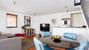 En moderne stue med en grå sofa, et glasbord med blå stole, et fladskærms-tv og et køkkenområde med et hvidt skab og en lille plante.