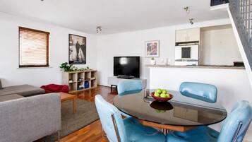 A modern living room with a grey sofa, a glass dining table with blue chairs, a flat-screen TV, and a kitchen area with a white cabinet and a small plant.